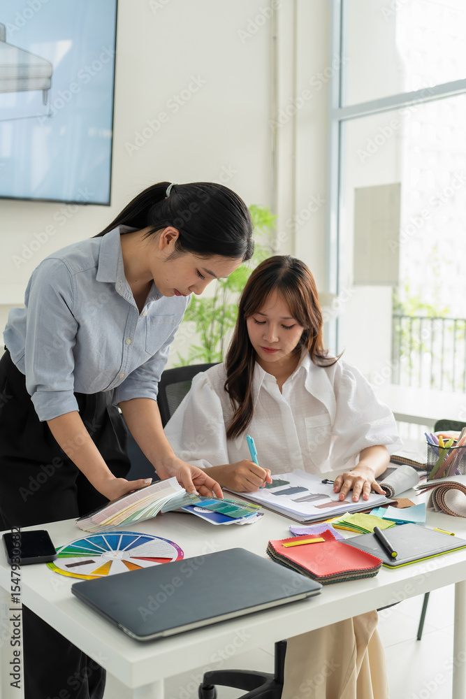 Fototapeta premium Two Asian women working together in a bright, modern studio.while using a laptop and smartphone. The creative workspace is filled with colorful samples for seasonal palette selection.