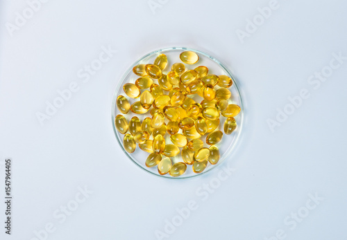 Top-view of yellow and amber supplement capsules in a glass petri dish, isolated on a white background. This shows scientific research, pharmaceutical development for health products like fish oi.
