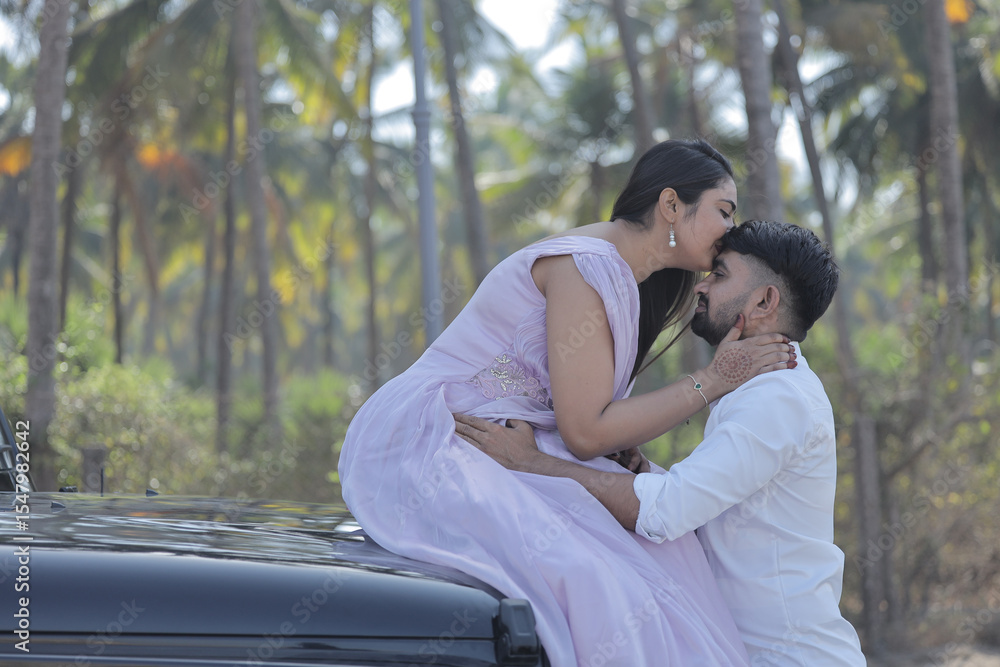 Naklejka premium A cheerful Indian couple shares a romantic moment on a palm-lined road, standing in front of a black SUV. The woman is wearing a flowing lavender gown, and the man embraces her from behind with a joyf
