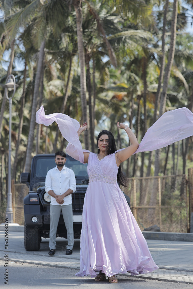 Obraz premium A cheerful Indian couple shares a romantic moment on a palm-lined road, standing in front of a black SUV. The woman is wearing a flowing lavender gown, and the man embraces her from behind with a joyf