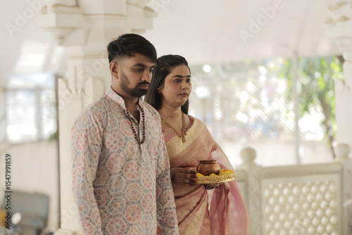 An Indian couple performs the sacred Abhishekam ritual to a Shiva Lingam in a Hindu temple. The woman is dressed in an elegant silk saree with jasmine flowers in her hair, and the man wears traditiona