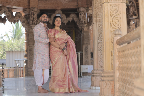 An Indian couple poses gracefully at a beautifully carved Hindu temple. The woman is wearing a rich golden-pink silk saree adorned with jewelry and bangles, while the man is dressed in an elegant ethn