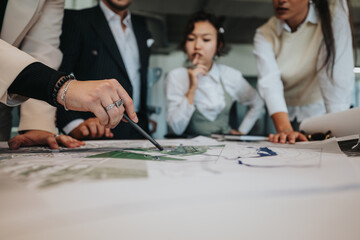 A group of diverse people gather around a table, analyzing blueprints. The project leader uses crutches while guiding the team's discussion on the project details.