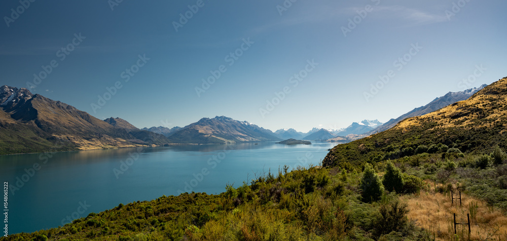 Fototapeta premium A breathtaking alpine lake stretches beneath snow-dusted peaks and golden hills, inviting stillness and wonder in a vast, painterly panorama of untouched natural harmony and distant mystery.