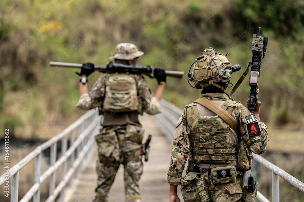 Fototapeta premium Two armed special forces soldiers in full tactical gear patrolling a narrow bridge during field training mission. Focused expressions, strategic movement in rural terrain.