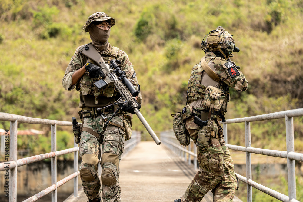 Fototapeta premium Two armed special forces soldiers in full tactical gear patrolling a narrow bridge during field training mission. Focused expressions, strategic movement in rural terrain.