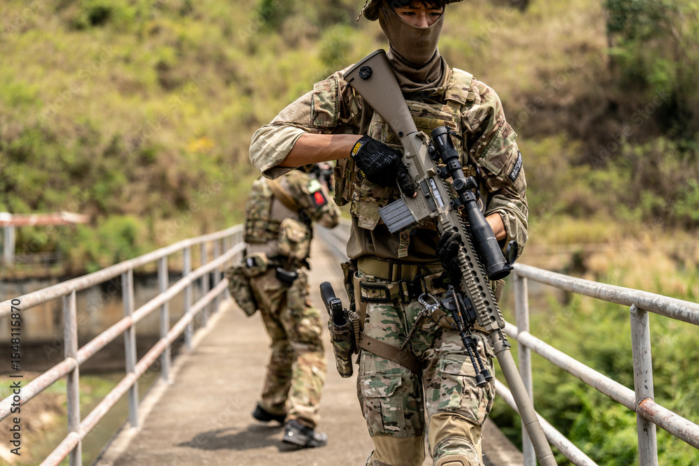 Fototapeta premium Two armed special forces soldiers in full tactical gear patrolling a narrow bridge during field training mission. Focused expressions, strategic movement in rural terrain.