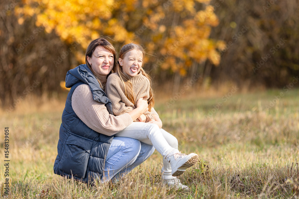 Fototapeta premium Loving mother hugging her little daughter during a peaceful autumn walk in the park, surrounded by vibrant fall foliage. Warmth and affection moment, capturing the beauty of family bonding. Copy space