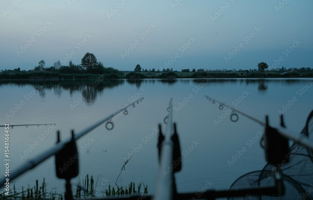 Obraz premium Fishing rods with bite alarms set up by a tranquil body of water, hinting at a peaceful day of carp fishing.