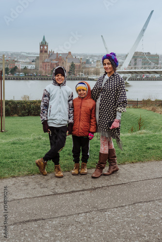 A mother and her young boys are posing in front of the modern Peace Bridge in Londonderry with the old walled city in the background