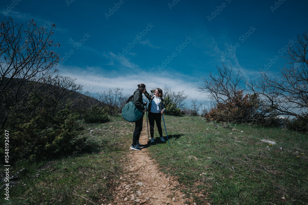 Obraz premium Two people with hiking gear and walking sticks explore a nature trail surrounded by green foliage and a clear blue sky, embracing the outdoor adventure on a sunny day.