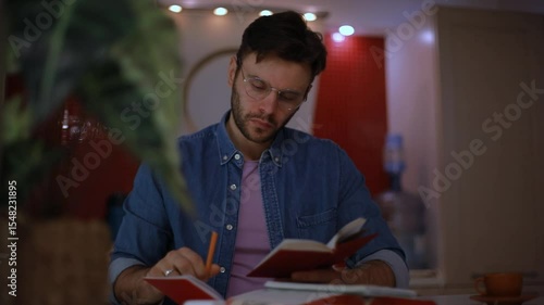 Focused young man in glasses writing in a notebook while reading, seated in a cozy kitchen. Evening scene with soft lighting, capturing study, planning, or journaling at home.
