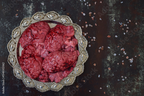 Beef brains arranged on ornate metal plate surrounded by salt grains over black textured surface under dim light. Halloween culinary atmosphere with eerie food styling and experimental setting.