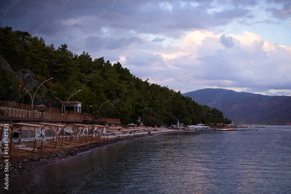 Fototapeta premium Serene Beachfront Overlooking Forested Hills Under a Dramatic Cloudy Sky