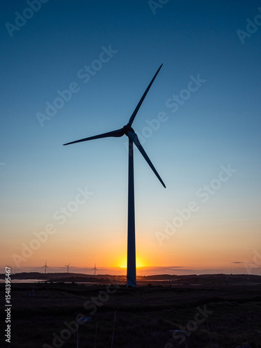 фотографія Silhouette of a tall wind turbine against blue sky and warm sun at the bottom of the mast
