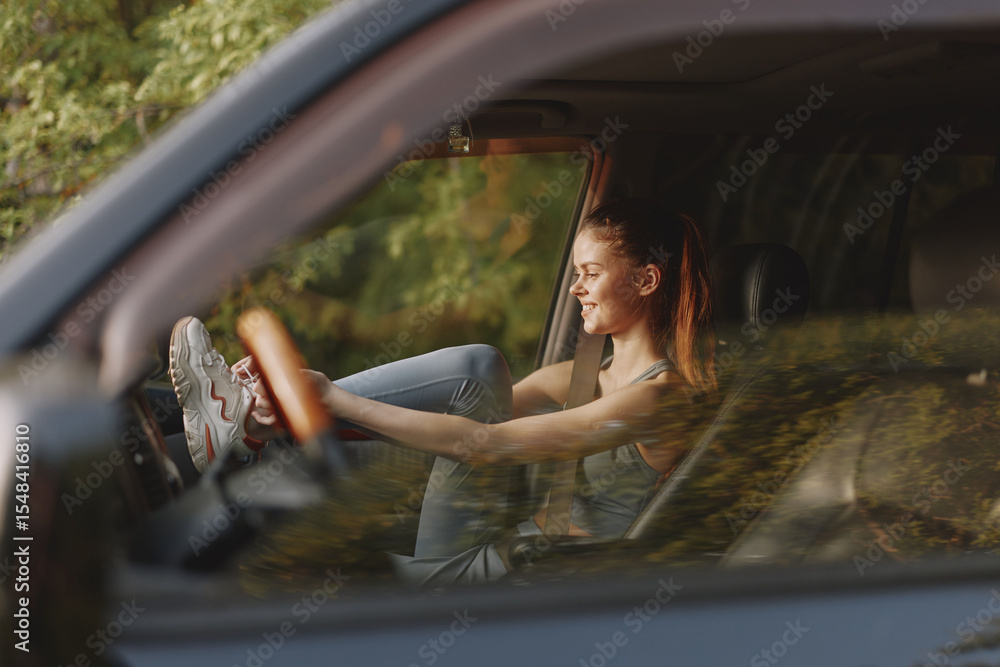 Fototapeta premium Happy young woman enjoying a road trip, smiling in car, wearing casual workout clothes, with greenery in the background, evoking a joyful travel spirit