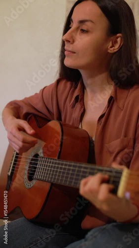 Smiling young woman playing acoustic guitar at home in a cozy room with warm lighting. She looks content and relaxed, enjoying a peaceful moment of creativity and musical expression.
