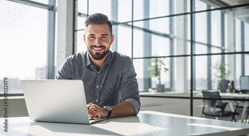 Smiling man working on laptop in bright office with large windows and modern interior design behind him