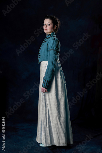A Regency woman wearing a blue cotton dress and a short spencer with soutache braid standing against a studio backdrop