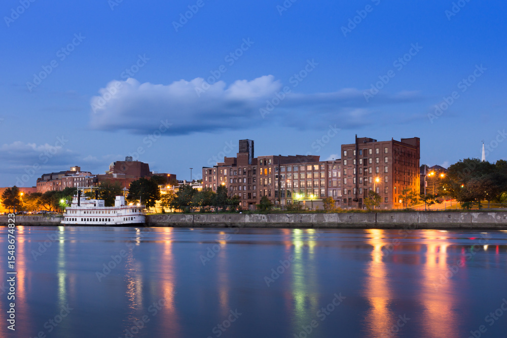 Obraz premium Long exposure panorama of Troy, NY at night from across the Hudson River with paddle wheeler, reflections, and lighted warehouses.