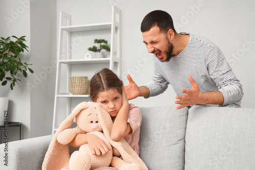 Scared girl with toy sitting on sofa and her angry father at home. Domestic v...