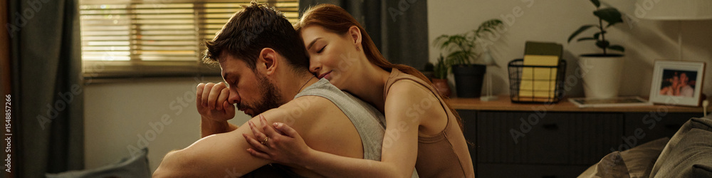 Fototapeta premium Man sitting on bed holding head in hand while woman embracing him from behind showing support during family problems