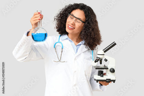 Female African-American scientist with microscope and flask on light background