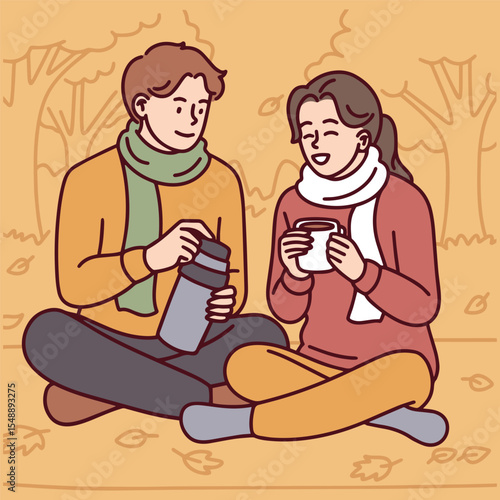 Couple in warm clothes sits on ground among fallen leaves, pouring hot tea from thermos. Peaceful autumn moment with cozy atmosphere