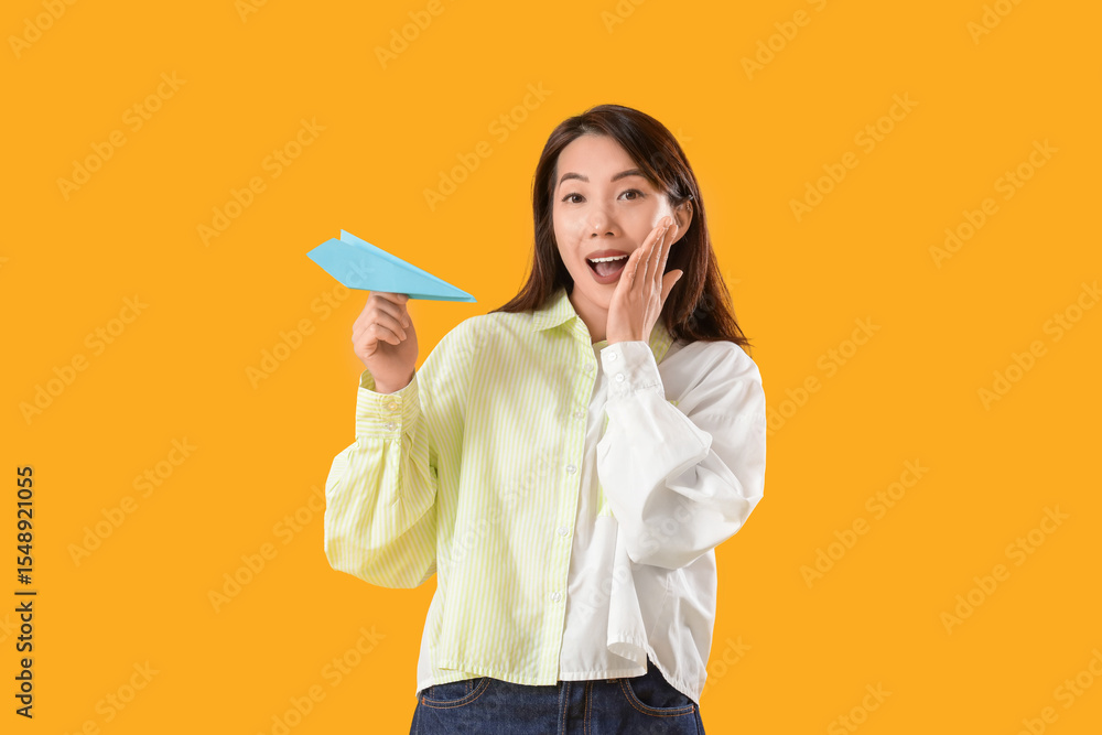 Surprised young Asian woman with paper plane on yellow background Surprised young Asian woman with paper plane on yellow background