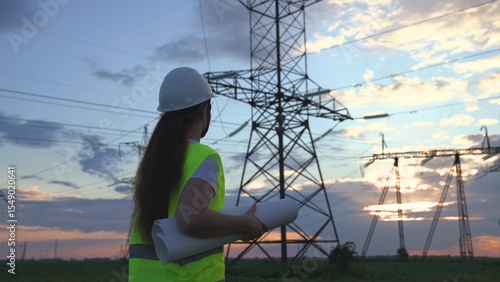 An electrician on the background of high towers of power plants looks at the project for the development of an electrical structure, the expansion of the electrical voltage of volts in the wires.