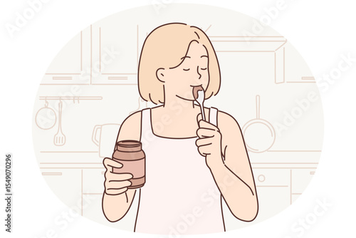 Happy woman stands in kitchen with jar of chocolate paste or peanut butter and licks spoon