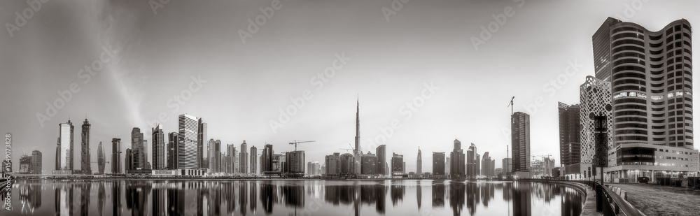 Fototapeta premium Monochrome city skyline with tall skyscrapers reflecting on calm water under a cloudy sky, Dubai Marina bay UAE