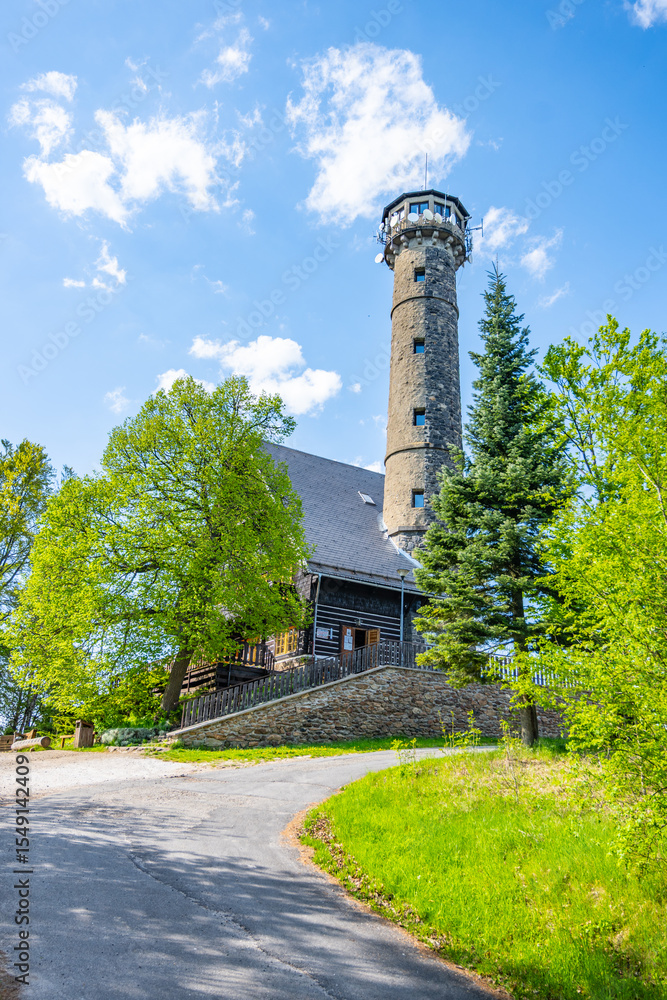 Fototapeta premium Svatobor lookout tower stands tall against a blue sky, surrounded by lush green trees. A well-maintained path leads to this popular tourist attraction in the serene countryside.