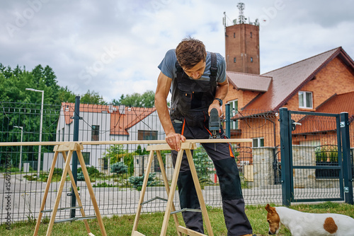 Man using electric jigsaw to cut wooden plank outside near residential buildings. Outdoor carpentry and DIY work and home improvement concept