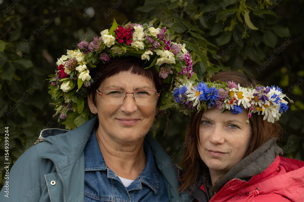 Obraz premium Two women stand close together wearing colorful floral wreaths made of wildflowers and greenery, smiling gently against a leafy natural backdrop.