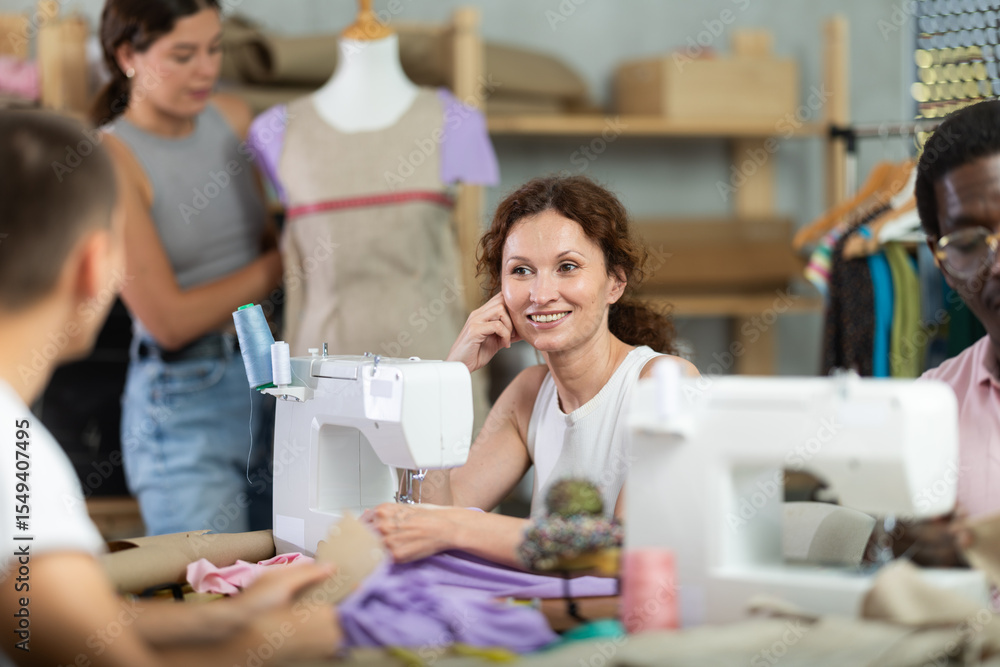 Obraz premium Cheerful woman working at sewing machine and friendly chatting with participants collaborating, fitting garment and preparing fabric during creative course on fashion design for adults