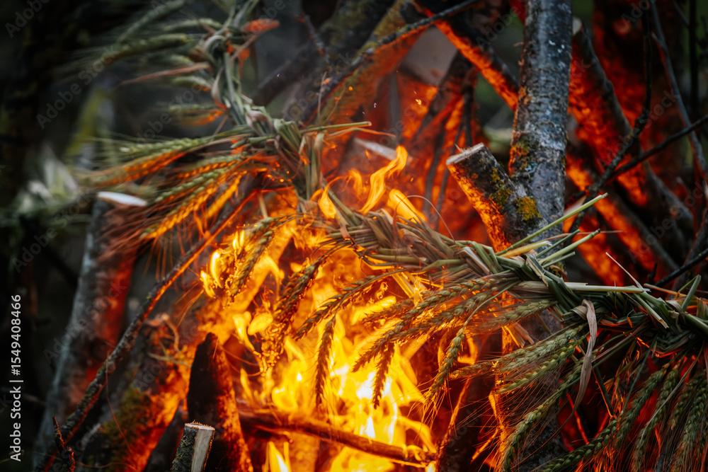Close-up of a traditional braided wheat ornament burning in vivid orange flames, surrounded by branches and intense firelight. Close-up of a traditional braided wheat ornament burning in vivid orange flames, surrounded by branches and intense firelight.
