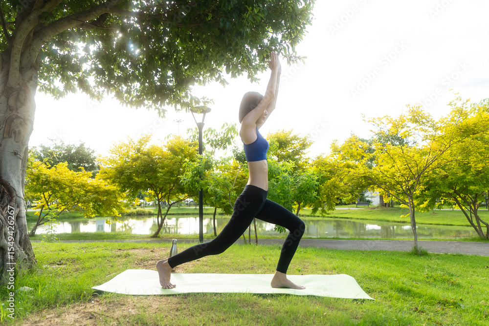 Fototapeta premium Young asian woman in sportswear stretching on a gym mat with a water bottle in a park, doing yoga. The young woman is taking care of her physical and mental health, wellbeing concept.
