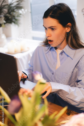 Young shocked businesswoman annoyed unpleasant spam message, open mouth, unexpected news, looking at laptop screen while sitting and working from home.