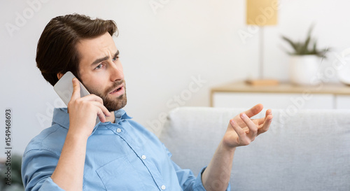 Foto Discontented Guy Frowning Talking On Cellphone Sitting With Laptop, Having Computer Or Internet Connection Problem At Home