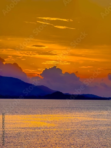 A dramatic sunset paints the sky in fiery oranges and yellows, with soft purple and blue clouds hovering above distant, silhouetted mountains. The vast expanse of the ocean reflects the vibrant hues