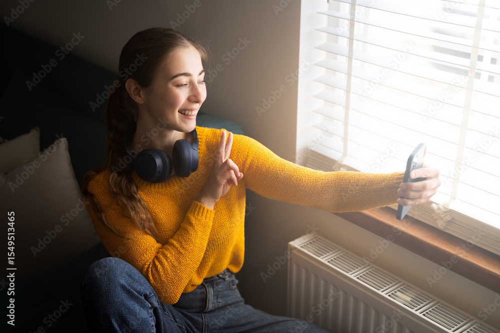Fototapeta premium Young woman smiling during video call on smartphone, wear headphones and casual clothes show peace sign sitting by window in living room, virtual meeting at home, side view portrait