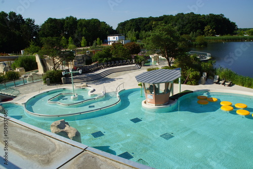 Beneath a radiant sky, a large public swimming pool stretches out, lined with relaxed swimmers and colorful lounge chairs. The sun sparkles on the water’s surface, creating shimmering reflections that