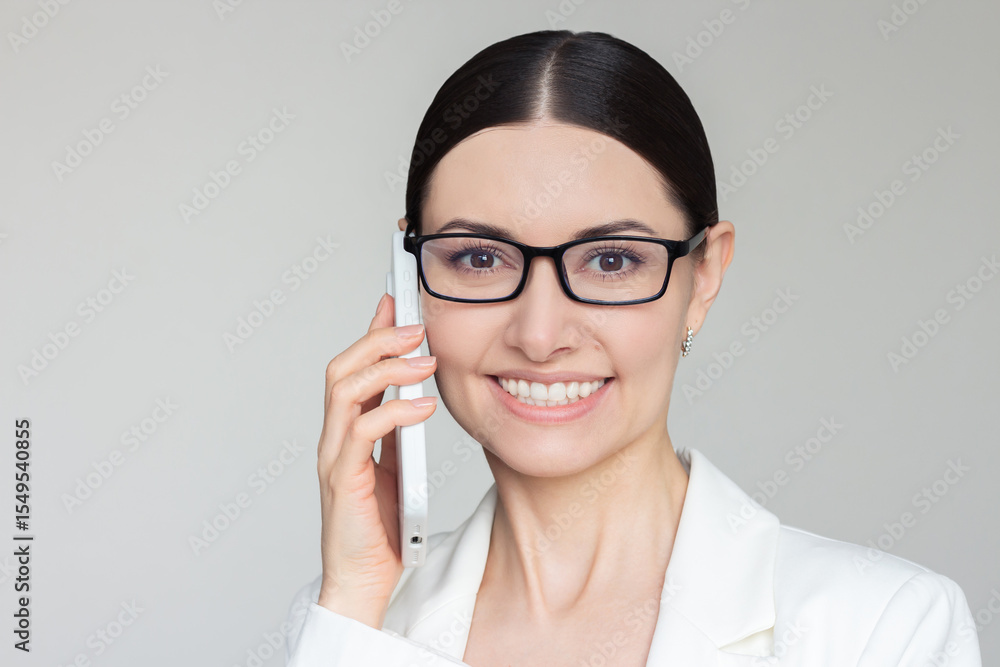 Young pretty Caucasian brunette businesswoman in glasses wearing white jacket talking on the smartphone looking at camera and smiling. Mobile communication. Young pretty Caucasian brunette businesswoman in glasses wearing white jacket talking on the smartphone looking at camera and smiling. Mobile communication.