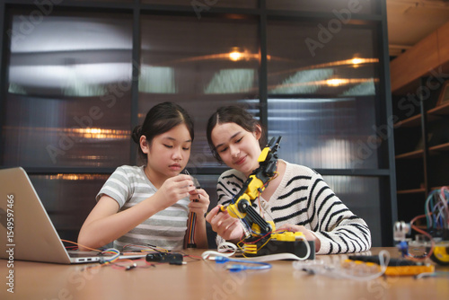 Two young Diverse girls concentrating on assembling electronic wires and robotic arm on table in classroom, actively learning STEM engineering and teamwork in a hands-on tech education project