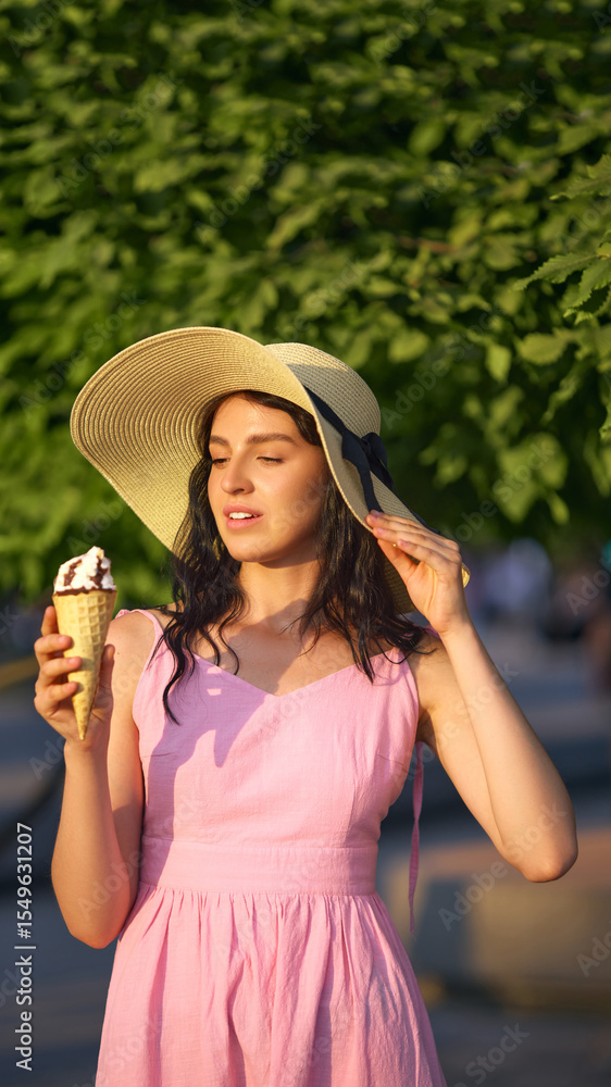Fototapeta premium A Caucasian woman enjoys an ice cream outdoors, wearing a sun hat and pink dress on a sunny day. Concept of summertime leisure and carefree relaxation. Casual fashion and summerwear
