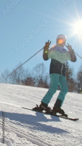 Female beginner skier posing show V symbol playfully love skiing on groomed snow slope under bright winter sun during ski vacation learning to ski alone. Intentional out of focus background