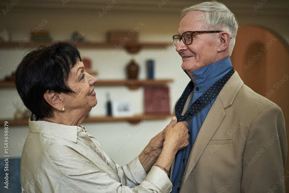 Obraz premium Elderly couple enjoying tender moment as woman helps man with tie, sharing genuine smiles. Background features bookshelf with various items adding cozy ambiance
