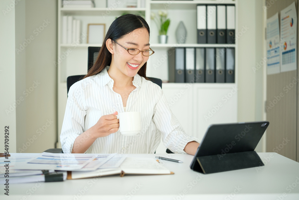 Asian businesswoman analyzing charts and graphs at her desk with a digital tablet and coffee mug in hand. Asian businesswoman analyzing charts and graphs at her desk with a digital tablet and coffee mug in hand.