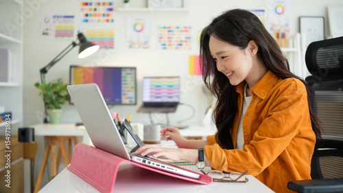 Smiling young female graphic designer working on a laptop in a  creative home office.